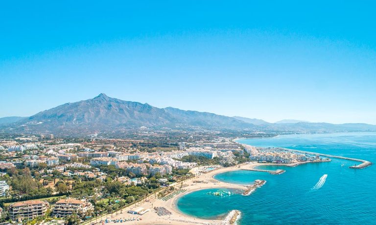 Aerial view of Marbella's coastline with clear blue skies, showcasing the vibrant cityscape against the backdrop of the iconic La Concha Mountain, embodying the prime real estate and investment potential of the region.