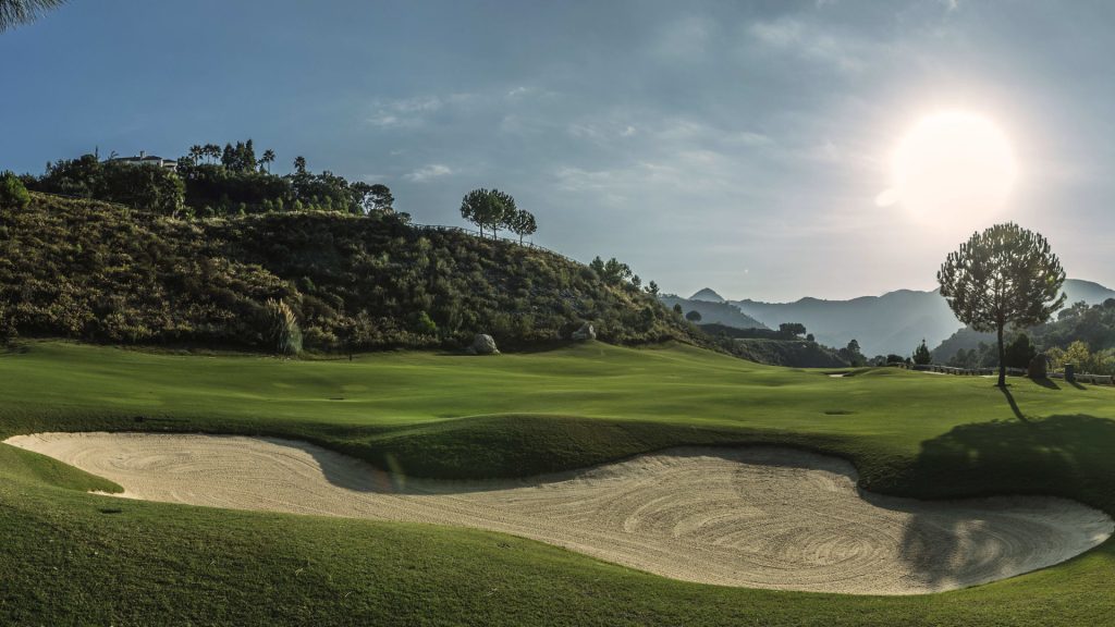 Early morning light casting shadows on the undulating greens and sand traps of a golf course in Marbella with mountains in the distance.