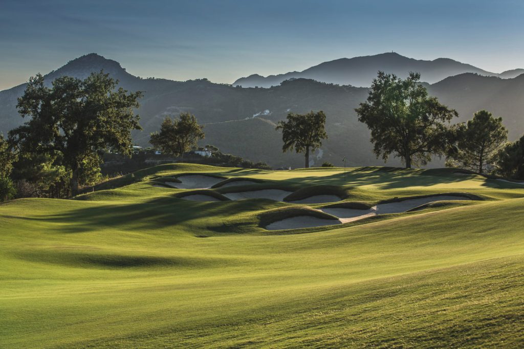 Early morning light casting shadows on the undulating greens and sand traps of a golf course in Marbella with mountains in the distance.