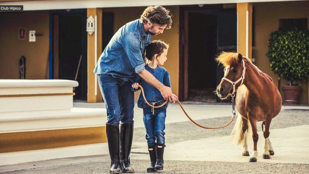 A father and child in matching denim outfits bonding over a pony at a Marbella equestrian club, under the warm Spanish sun.